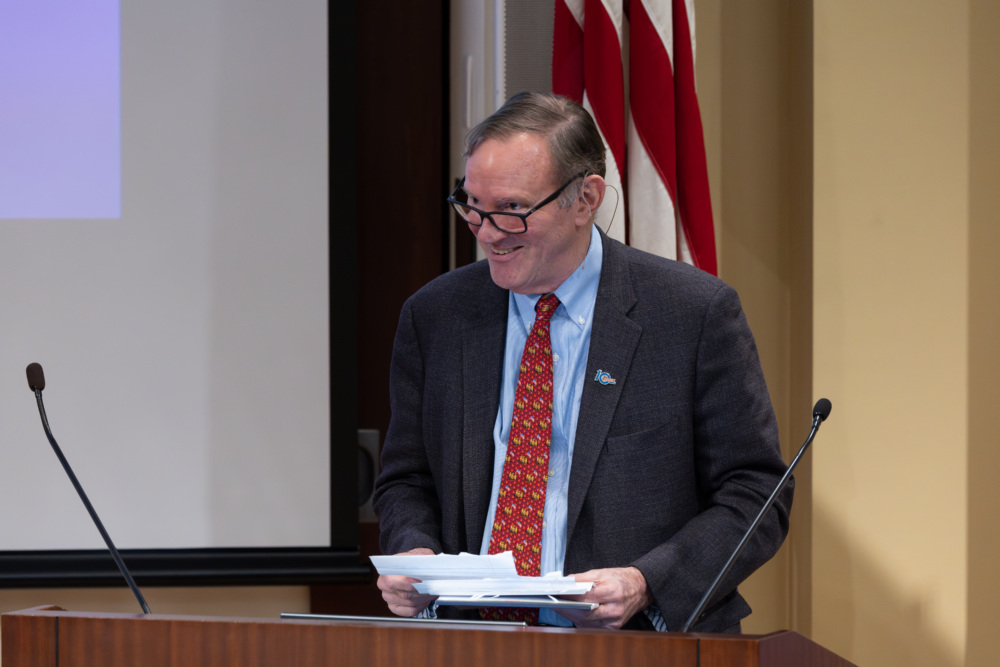 Don Graham smiles while standing at a podium at the Social Finance Institute launch event, holding papers and wearing a red tie. flags and a projection screen are visible in the background.